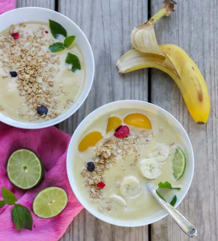 two white bowls filled with smoothies topped with bananas, peaches, mint and granola on wooden surface with pink napkin, mint, limes and bananas