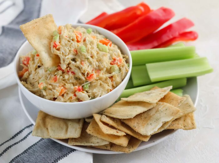 white and black napkin with plate of pita chips, celery, and red bell peppers, and a bowl of chicken salad with pita chip in bowl