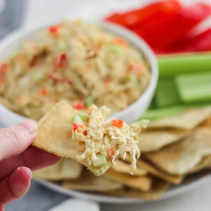hand holding a pita chip with hummus chicken salad on it, background has bowl and sliced veggies