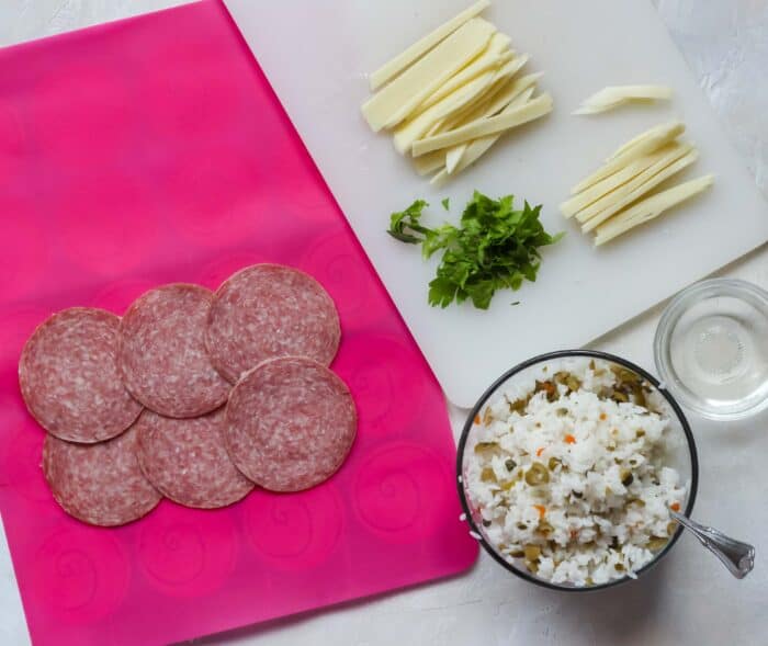beef salami on pink baking liner, rice with capers and olives, celery leaves, sliced cheese on cutting board