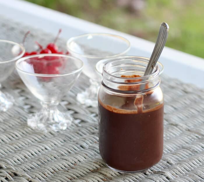 jar of chocolate sauce with a spoon in it. empty small glass ice cream cups with a bowl of cherries on a wicker table