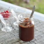 jar of chocolate sauce with a spoon in it. empty small glass ice cream cups with a bowl of cherries on a wicker table