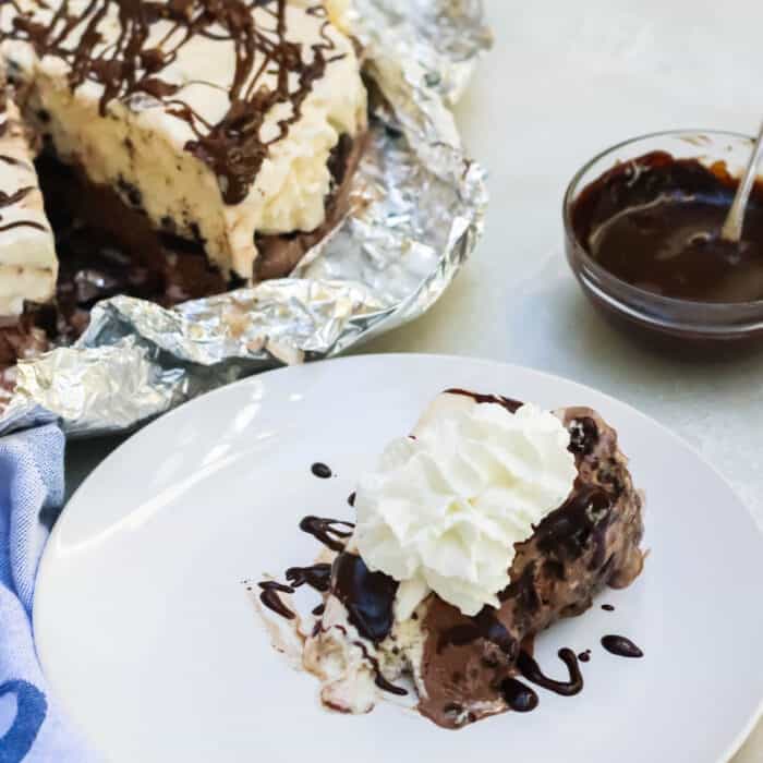 white plate with a slice of ice cream cake covered in whipped cream and chocolate sauce with ice cream pie in background