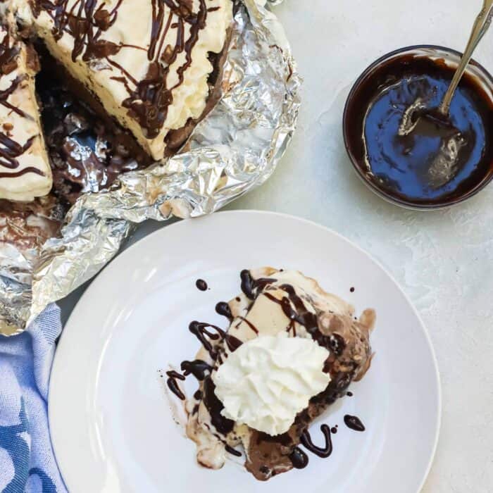 bowl of chocolate sauce with silver spoon, ice cream cake on foil and a plate with a slice of ice cream pie topped in whipped cream