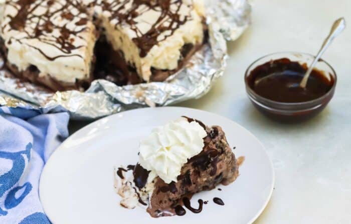 white plate with slice of ice cream cake covered in chocolate sauce and whipped cream with a bowl of chocolate sauce and ice cream cake in background