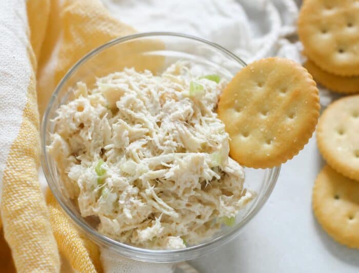 glass bowl of chicken salad with a butter cracker scooping into chicken salad on white table with a yellow and white towel