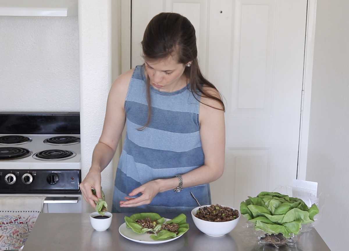 brunette female dipping a lettuce wrap into sauce while standing in the kitchen. extra lettuce and ingredients are on the table