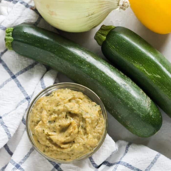 small glass bowl of zucchini sauce on a white and blue napkin with fresh zucchini, onion and lemon