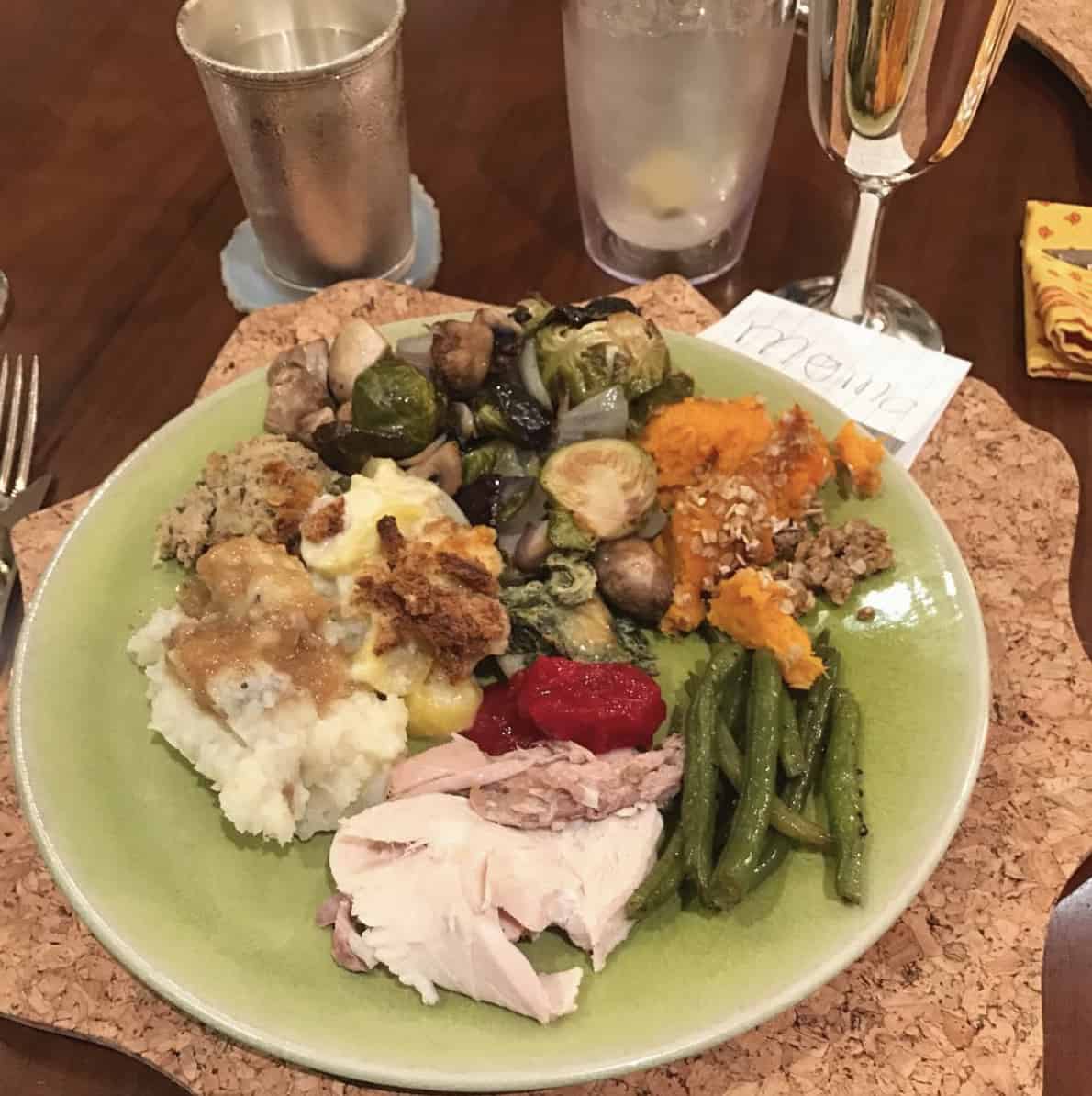 green plate with turkey, cranberry sauce, mashed potatoes and gravy, roasted thanksgiving veggies, sweet potato casserole and sauteed green beans on a table with wine and water glasses