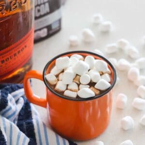 orange mug of hot chocolate with marshmallows and bottle of bourbon with container of cocoa powder on blue and white towel