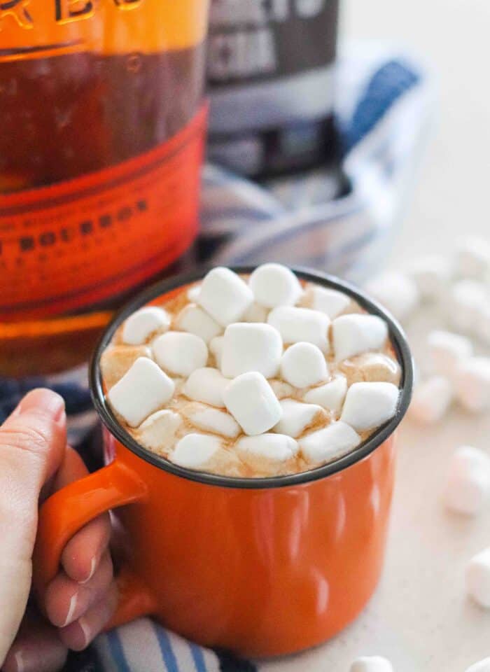 hand holding orange mug of hot chocolate topped with marshmallows and a bottle of bourbon with cocoa powder