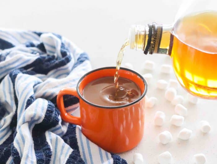 bourbon pouring into an orange mug of hot chocolate on a blue and white towel with marshmallows
