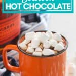 orange mug with hot chocolate and marshmallows on blue and white towel with bottle of bourbon and cocoa powder