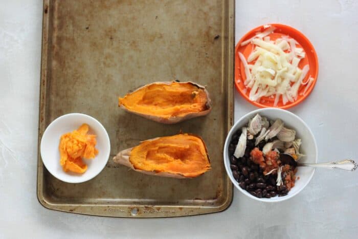 baked sweet potato on a baking sheet with some scooped out into a white bowl, a white bowl with salsa, beans, and chicken, and an orange top with shredded cheese