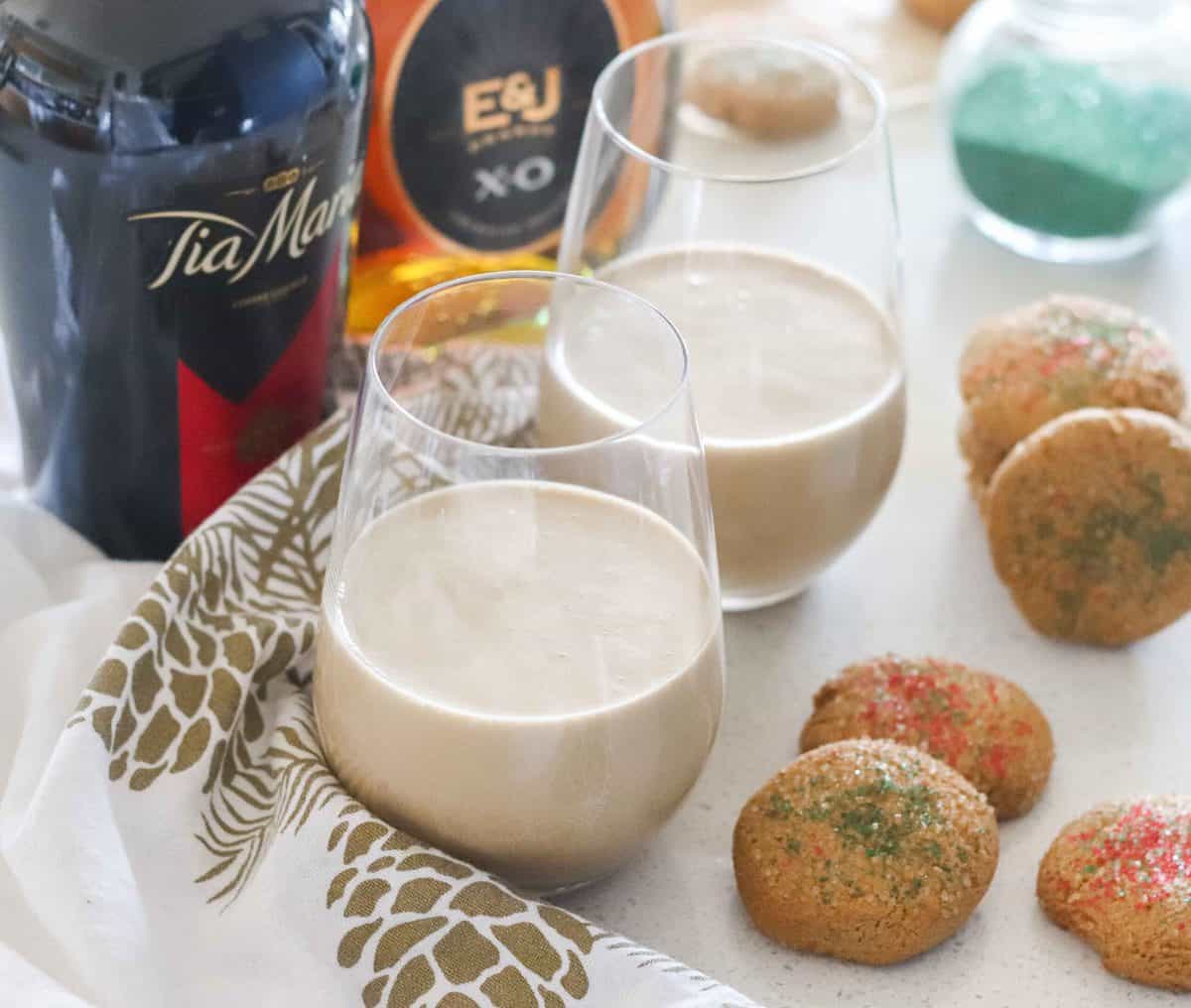two cream-colored cocktails in wine glasses on white and gold napkin next to cookies and bottles of brandy and coffee liqueur