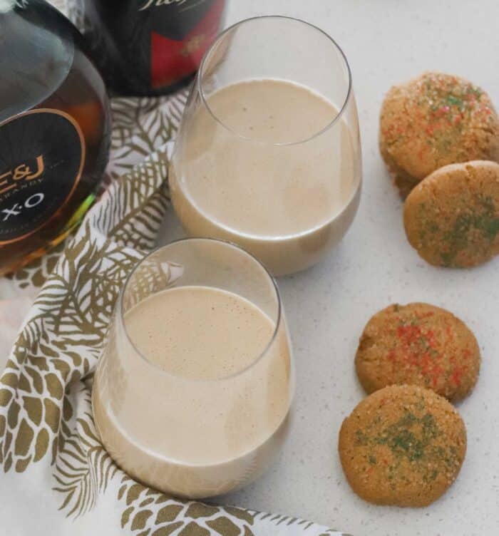 two wine glasses with cream colored coffee drinks on white counter with white and gold napkins plus Christmas cookies with green and red sprinkles and bottles of alcohol