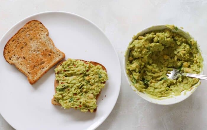white plate with two slices of toast with one slice topped with avocado and a white bowl of mashed chickpeas and avocado with a fork in the bowl