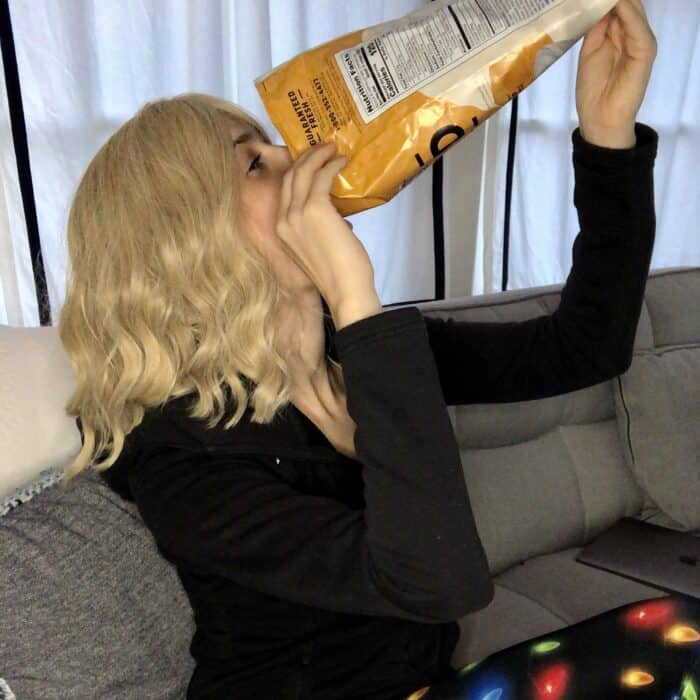 blonde female sitting on a grey sofa pouring chips into her mouth