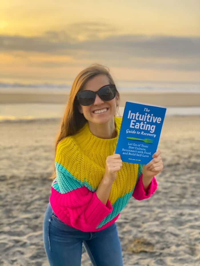 Meme Inge on a beach holding her book titled The Intuitive Eating Guide to Recovery