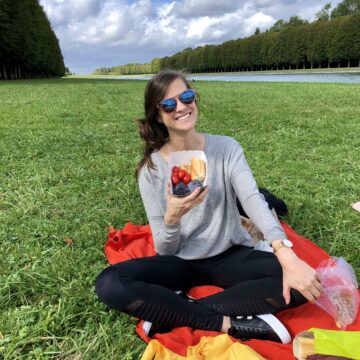 brunette female sitting in a blanket in Versaille garden smiling and holding a container of container of food she is about to be eating