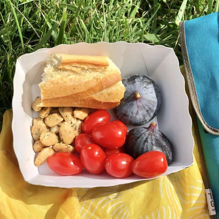 paper container with fresh figs, cherry tomatoes, marcona almonds, and a big piece of french bread sitting on a yellow napkin in the grass