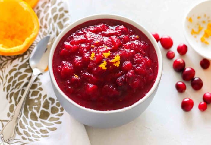 grey bowl with cranberry sauce topped with orange zest on a white counter with orange peel, white and gold napkin, and fresh cranberries