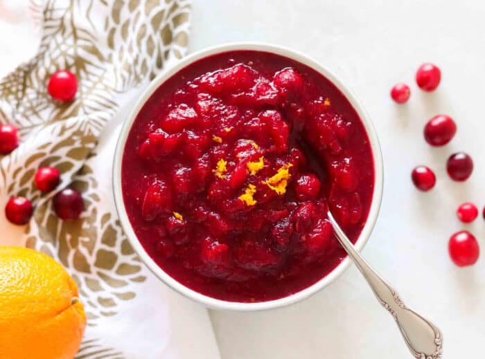 bowl of cranberry sauce with silver spoon on white counter with fresh cranberries, orange, and a white and gold napkin