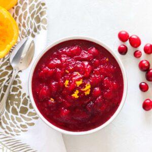 white counter with fresh cranberries, gold and white napkin, silver spoon, orange peel, and a bowl of cranberry sauce topped with orange zest