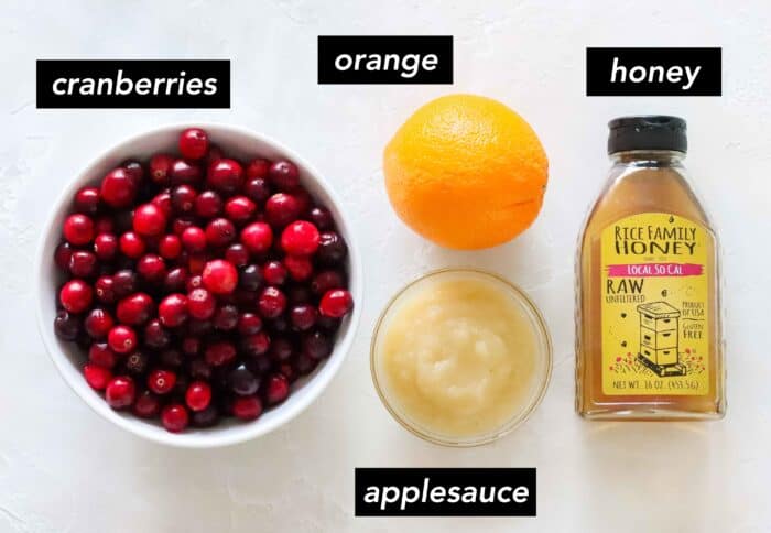 white bowl with cranberries, orange, clear bowl of applesauce, bottle of honey on white counter with text overlay describing ingredients