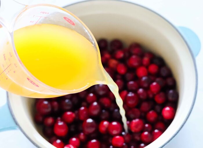 measuring cup of orange juice pouring into a saucepan full of cranberries