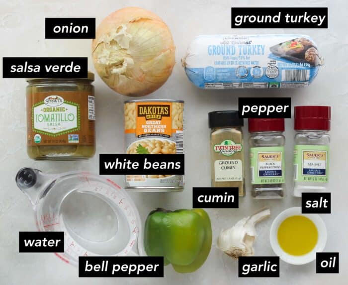 onion, tomatillo salsa, measuring cup of water, green bell pepper, can of white beans, ground turkey, cumin, pepper, salt, small bowl of oil, and a head of garlic