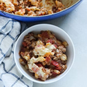 white bowl with hominy, tomatoes, beef, and cheese on a white and blue checkered napkin with a blue pot in background