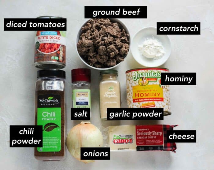 canned tomatoes, container of chili powder, salt grinder, onion, black of cheese, jar of garlic powder, can of hominy, small bowl of cornstarch, bowl of ground beef