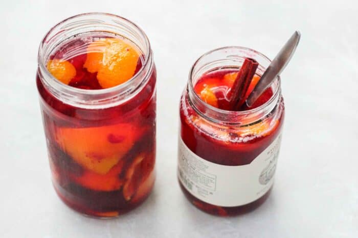 two jars full of cranberry infused vodka with orange peels. one jar has a cinnamon stick and a silver spoon coming out of the opening