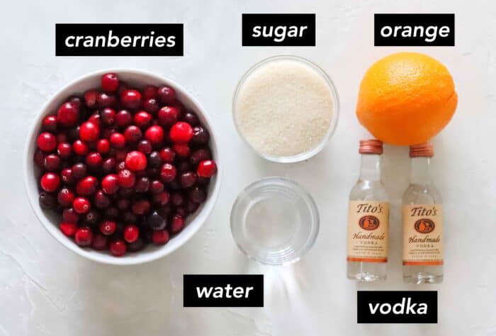 white counter with bowl of fresh cranberries, bowl of sugar, orange, two mini bottles of Tito's vodka, and a bowl of water with text overlay that describes each ingredient