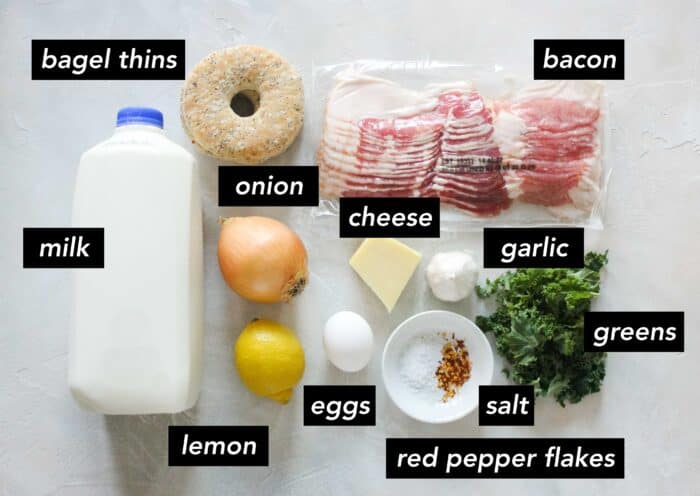 white counter with milk, bagel thin, package of bacon, onion, block of cheese, garlic, kale, bowl of salt and red pepper flakes, egg, and lemon