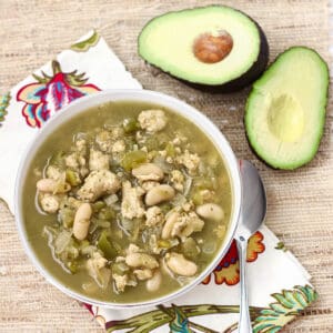 white bowl of white bean turkey chili with avocado and silver spoon on straw placemat with napkin