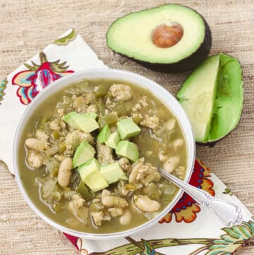 cut open avocado on a straw placemat with a white bowl of white turkey chili with a silver spoon on a patterned napkin