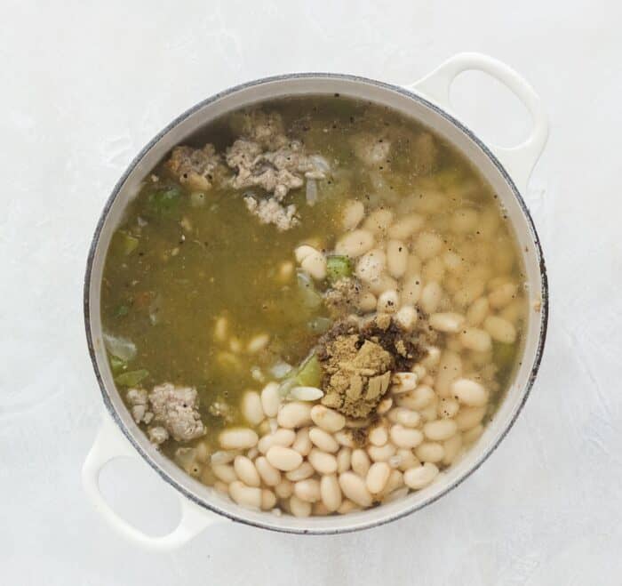 cream-colored pot on white counter with white beans, spices, bell peppers, onions, turkey, and broth