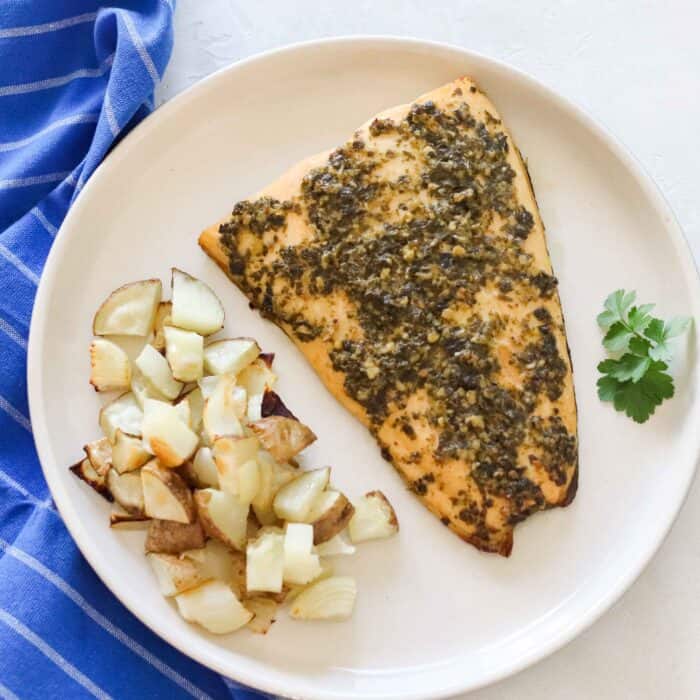 white plate with roasted potatoes and onions, parsley, and pesto salmon with a blue napkin