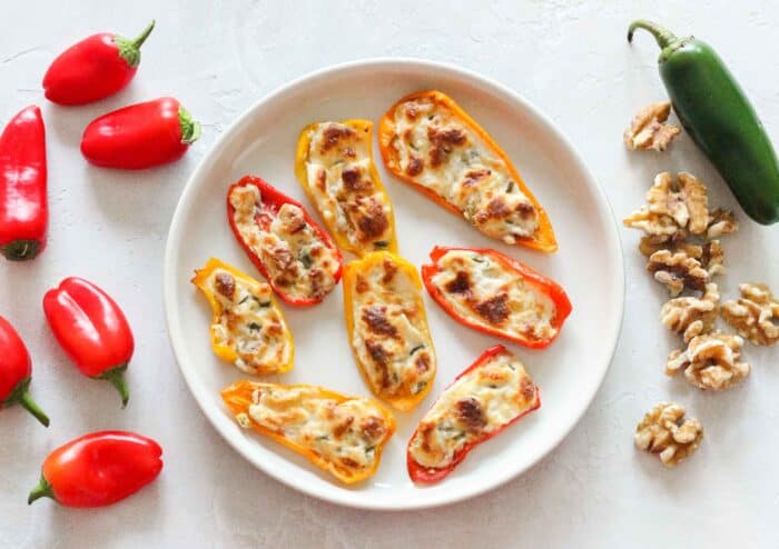 red mini bell peppers, a white plate with baked cream cheese stuffed bell peppers, a jalapeno, and walnuts