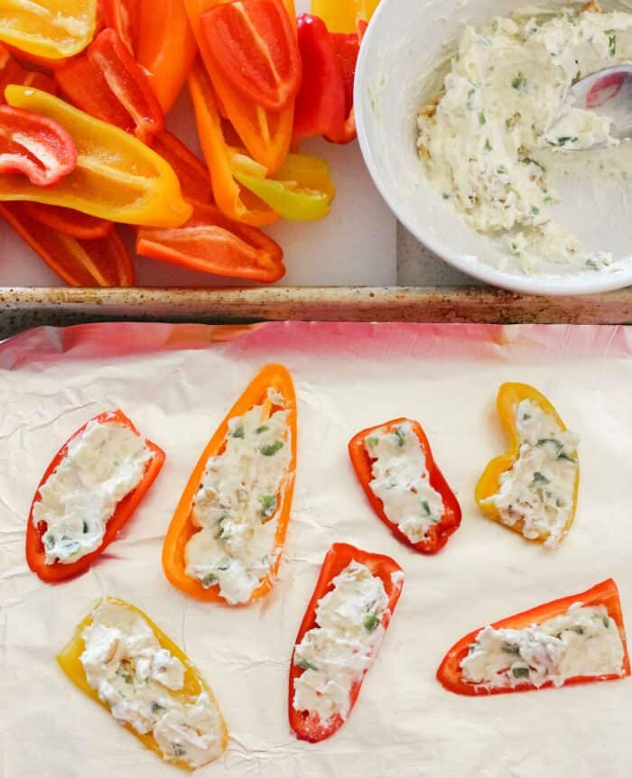 halved mini bell peppers, a bowl of cream cheese, and a foil lined baking sheet with cream cheese stuffed bell peppers