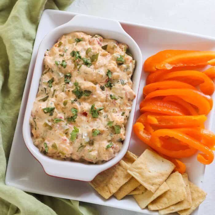 rectangle serving dish with crawfish dip on a square plate with sliced orange bell peppers and pita chips with a green napkin