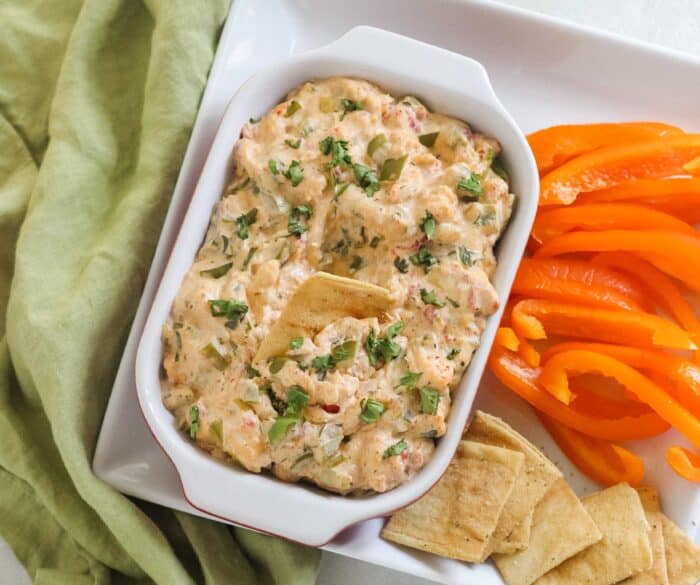 green napkin with square plate that has orange bell pepper slices, pita chips, and a rectangular serving dish filled with crawfish dip