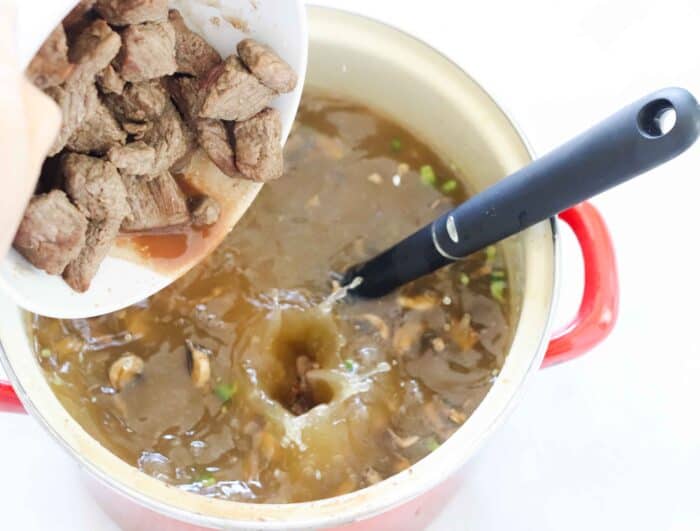 a hand pouring beef from a white bowl into a red soup pot with a broth based soup