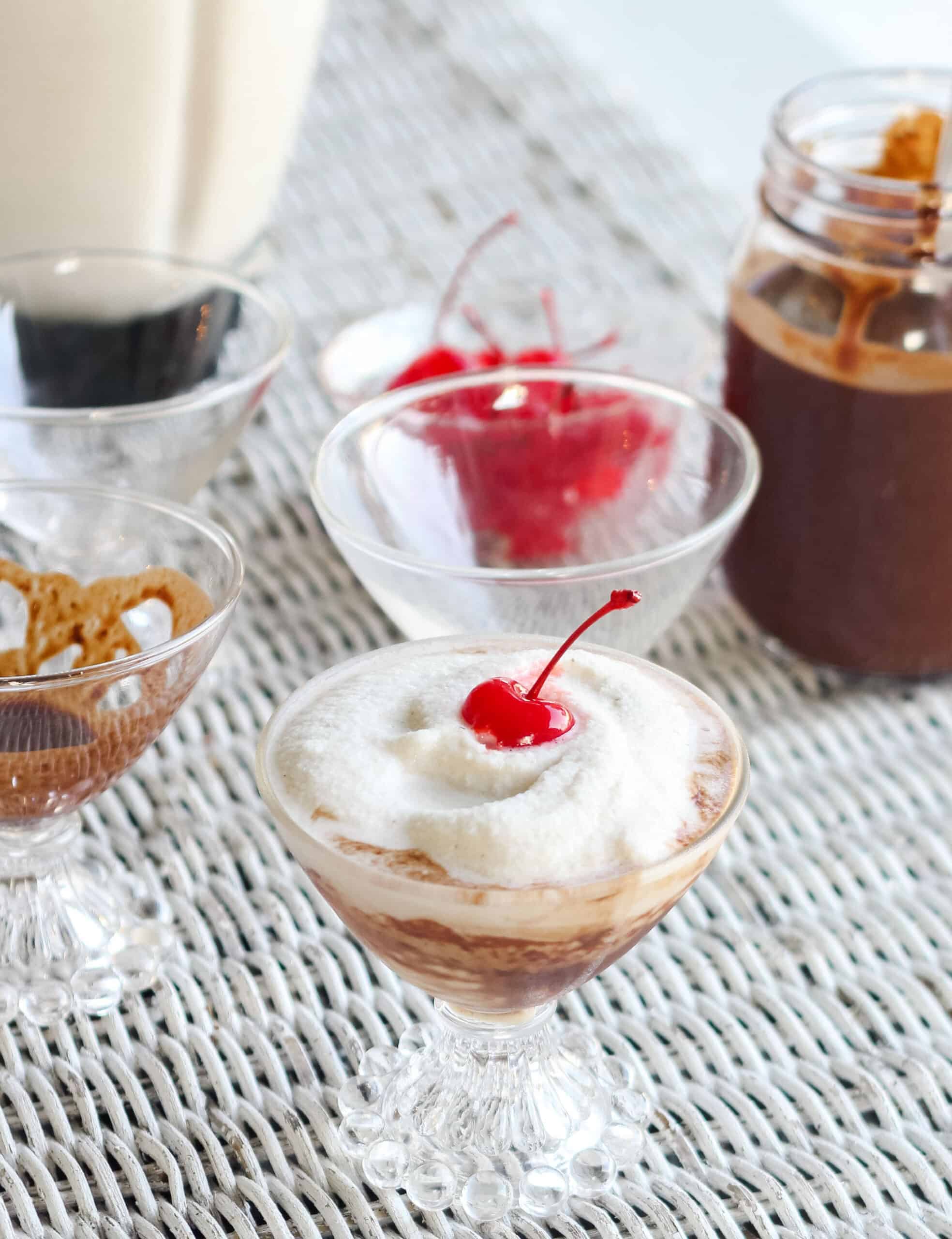 a blender filled with a frozen bushwacker, a glass with a bushwacker topped with a cherry and glasses with chocolate sauce