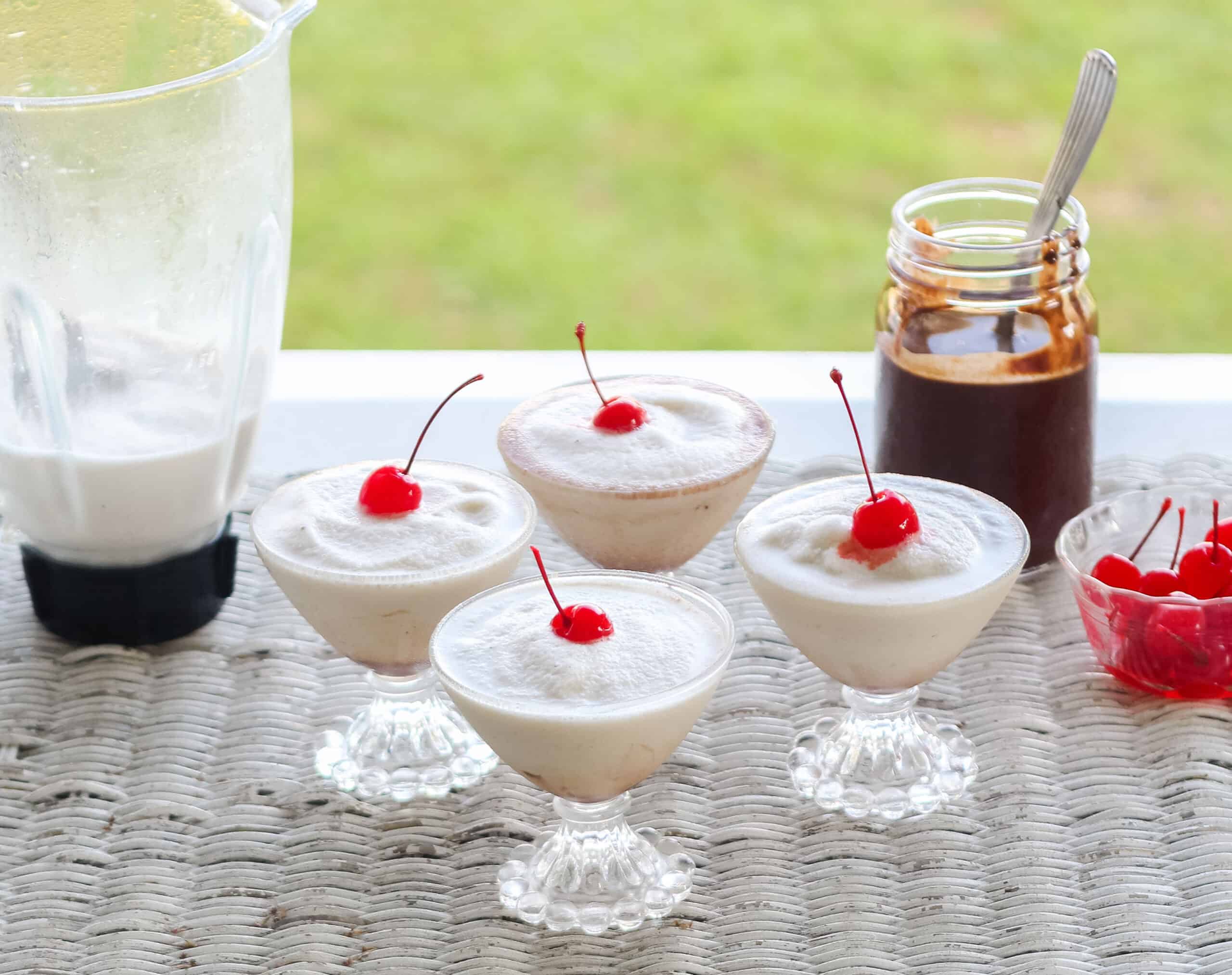 wicker table with blender full of frozen bushwacker drinks and four filled glasses with a jar of chocolate sauce on the side