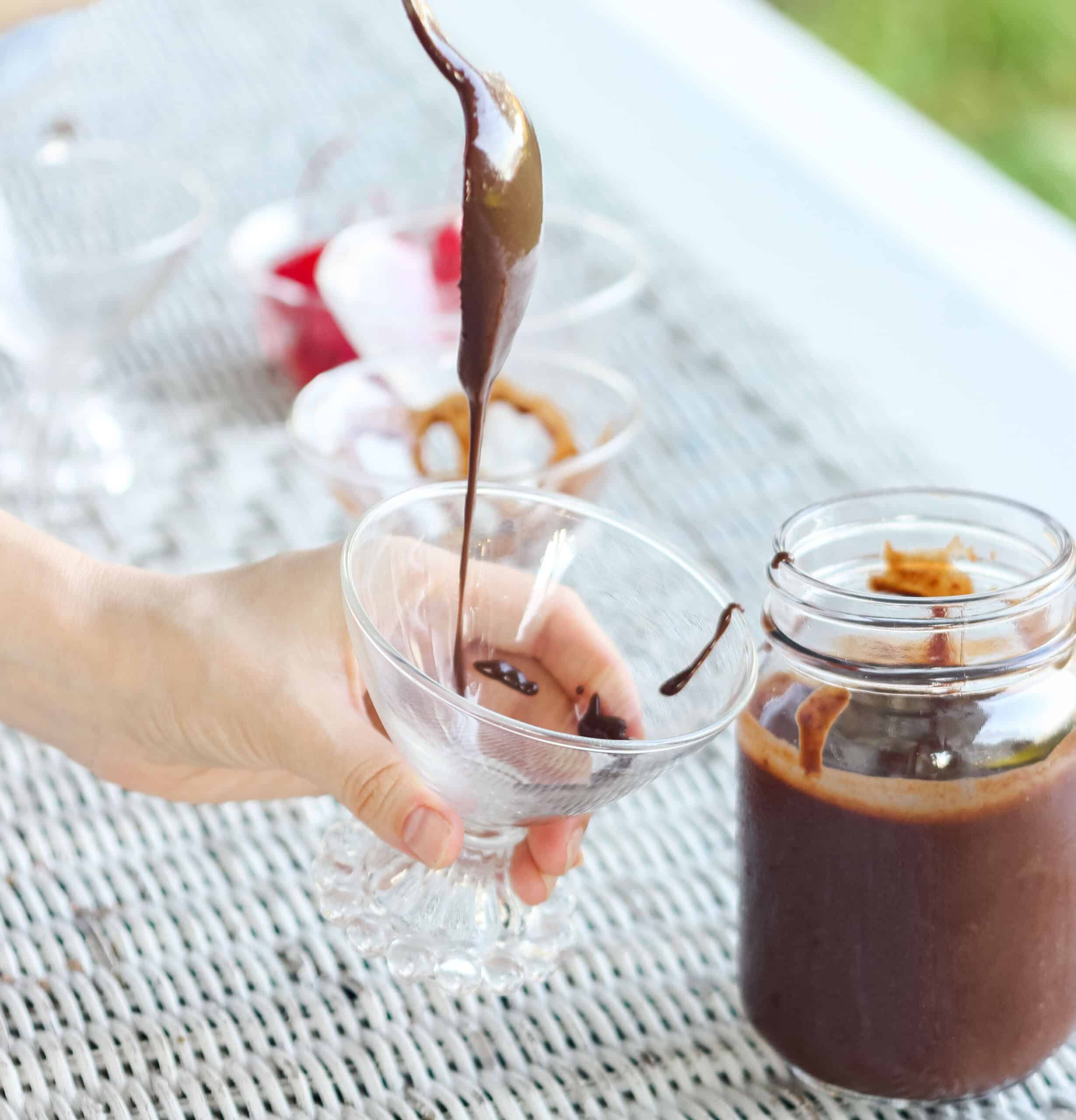 hand holding a glass with a spoon is drizzling chocolate sauce into the glass with a jar of chocolate sauce on the side