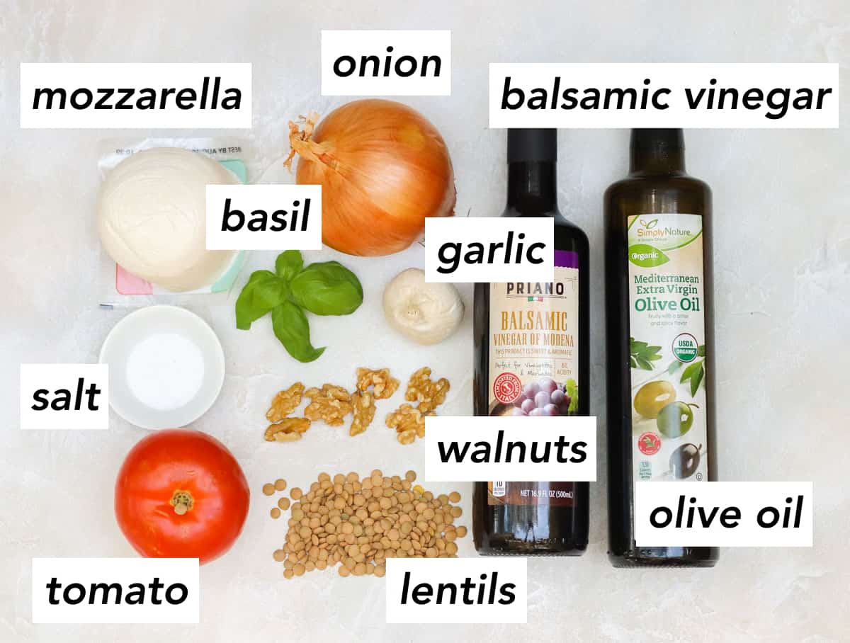 ball of fresh mozzarella, yellow onion, fresh basil, bowl of salt, large tomato, scattered lentils, scattered walnuts, head of garlic, bottle of balsamic vinegar, bottle of olive oil on a counter with text overlay describing each ingredient.