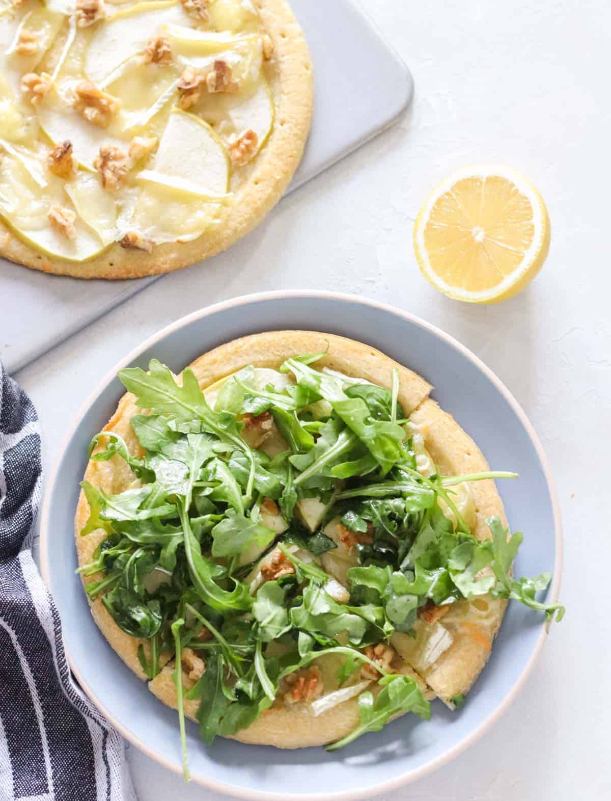 cutting board with brie apple pizza, plate with pizza topped with arugula, cut lemon
